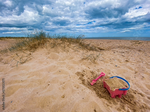 Pink Bucket and spade building a sand castle on the sand's hills on Great Yarmouth beach in a cold summer day, traditional UK, English East Coast, sky with clouds no people, large stretch of sand, UK 