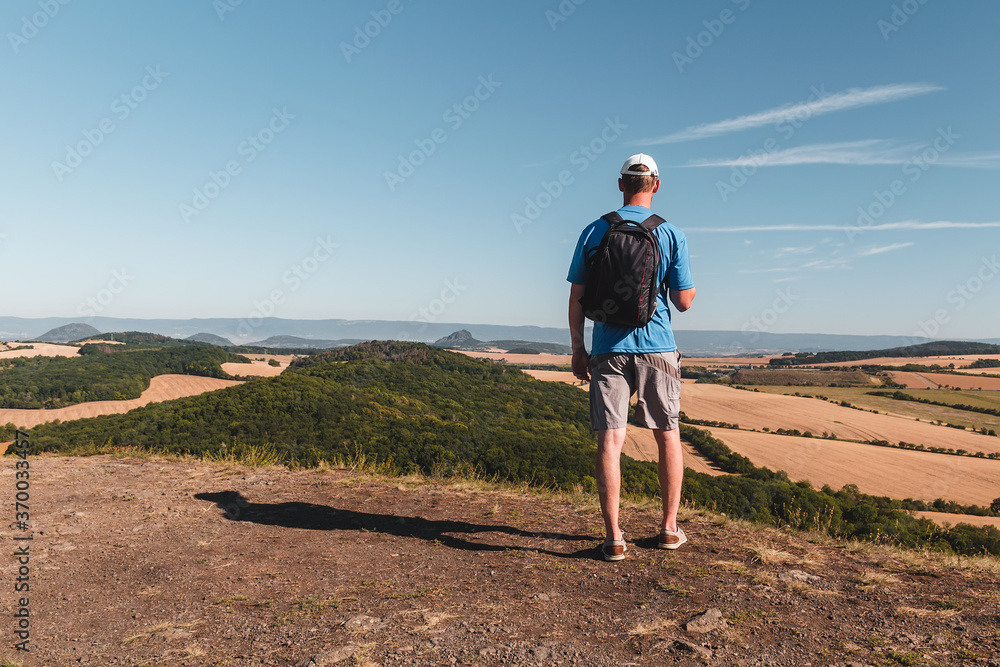 Young man with backpack standing on hill and looking to Czech central mountain valley at sunrise Young man with backpack standing on hill and looking to Czech central mountain valley at sunrise