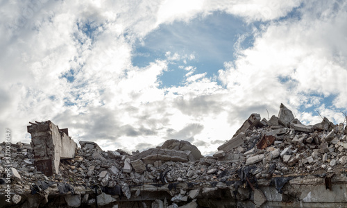 A pile of concrete gray debris of a destroyed building with a huge beam in the foreground against a blue sky with clouds