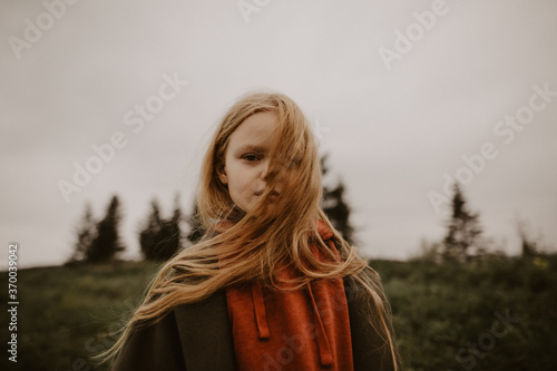 a little girl with blond hair looks seriously into the distance against the background of woods and houses
