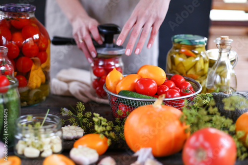 Woman's hands rolls glass preserves with jars sealer. Making homemade vegetables for coronavirus crisis. Tomatoes, pumpkins, garlic, peppers, pickles, zucchini lie on wooden table