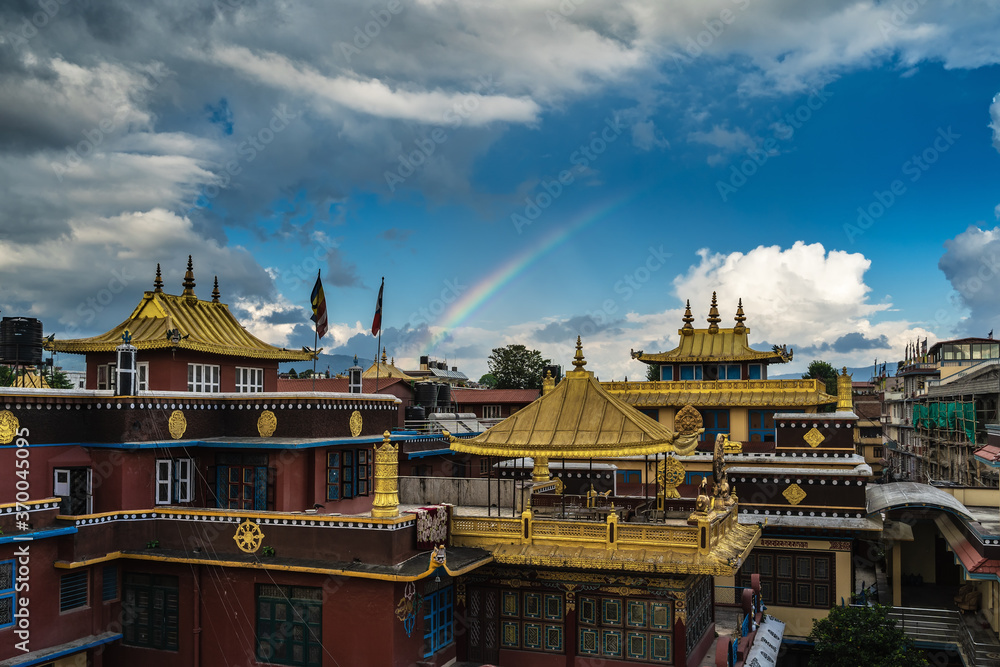 Fototapeta premium Bright rainbow arc over a Buddhist monastery with gilded roofs against the blue sky, dramatic gray clouds and mountains in the background.