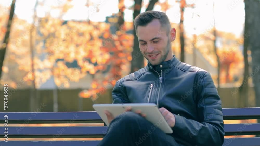 Student is Reading News or Watching Video on Tablet Computer in the Park in Autumn. Man in Black Leather Jacket is Sitting on a Banch Using Big White Tablet PC. Middle Close-Up Shot Student is Reading News or Watching Video on Tablet Computer in the Park in Autumn. Man in Black Leather Jacket is Sitting on a Banch Using Big White Tablet PC. Middle Close-Up Shot