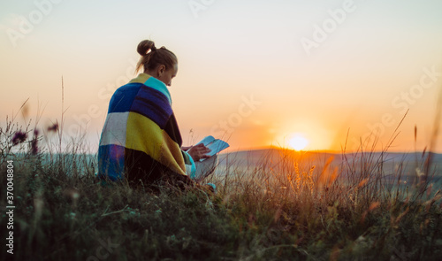 Back view of a young woman wrapped in a colourful knitted blanket seated on a hill at sunset, reading, meditating, contemplating