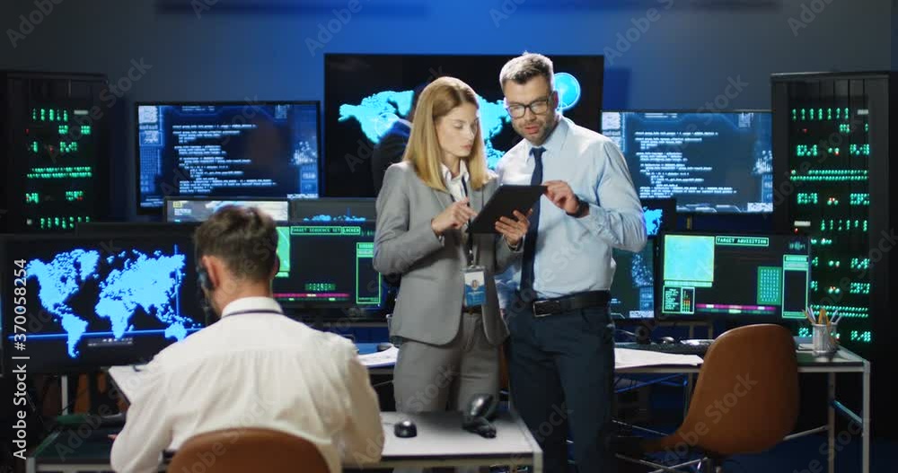 Caucasian male and female security information analytics talking about info on tablet device in monitoring governmental room. Professionals man and woman discussing digital surveillance in data center Caucasian male and female security information analytics talking about info on tablet device in monitoring governmental room. Professionals man and woman discussing digital surveillance in data center