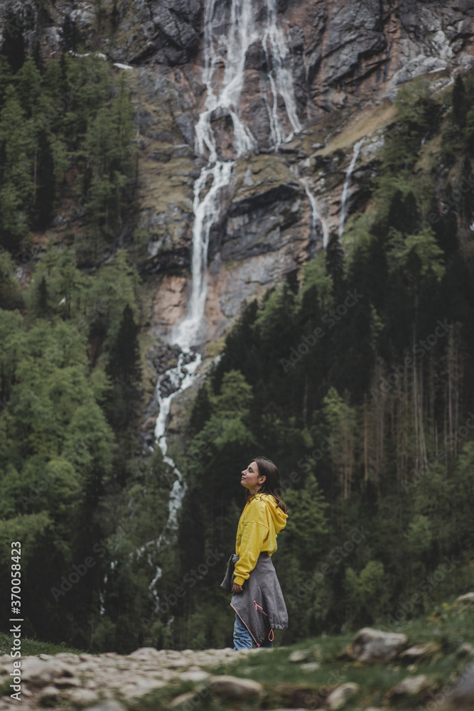 Young brunette girl in yellow sweatshirt closed eyes & feels freedom on background of Rothbach waterfall, pine forest & rock mountain cliff. Schonau am Konigssee. Bavaria. Germany