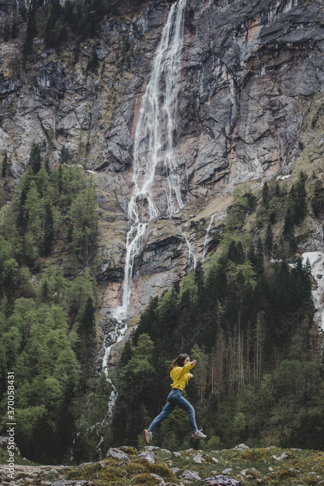 Obraz premium Young brunette girl in yellow sweatshirt jumps on background of Rothbach waterfall, pine forest & rock mountain cliff. Schonau am Konigssee. Bavaria. Germany