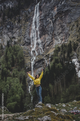 Schonau am Konigssee. Bavaria. Germany. Young caucasian girl in yellow sweatshirt raises her hands up & feels freedom on background of Rothbach waterfall, pine forest & rock mountain cliff