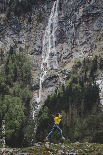 Young brunette girl in yellow sweatshirt jumps on background of Rothbach waterfall, pine forest & rock mountain cliff. Schonau am Konigssee. Bavaria. Germany