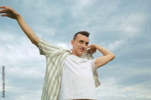 Handsome guy puts on a shirt against the background of the sky. Men's fashion, unity with nature.