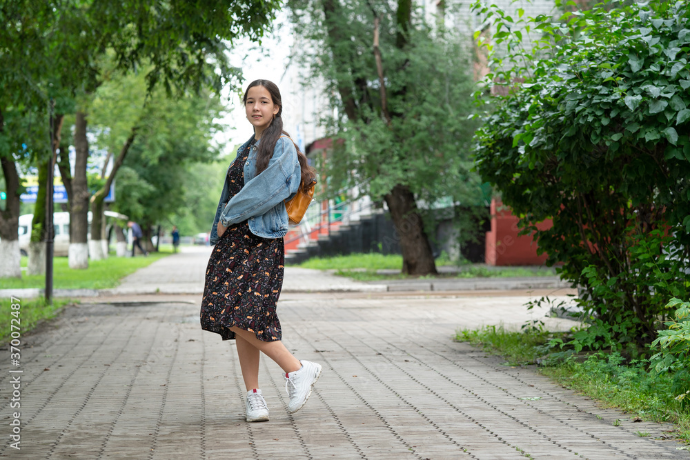 a girl in a dress and a denim jacket with an orange backpack on her shoulders goes to school and looks back. concept of back to school. Street photography. a girl in a dress and a denim jacket with an orange backpack on her shoulders goes to school and looks back. concept of back to school. Street photography.