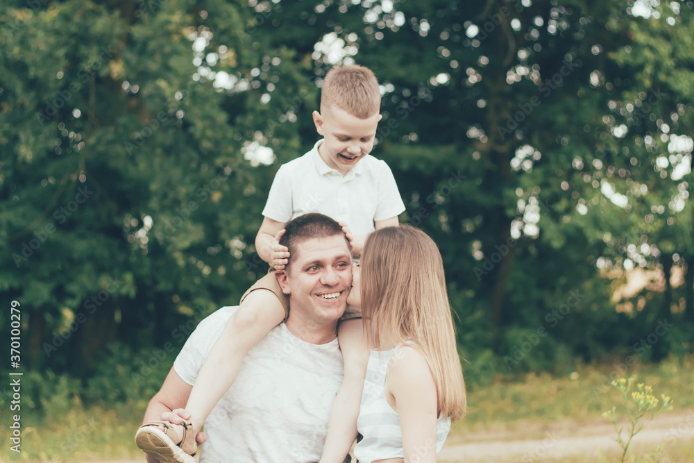 Family resting in nature, dad keeps the boy around his neck and mom kisses dad and smiles