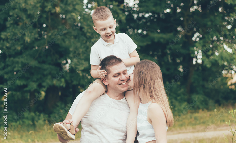 Family resting in nature, dad keeps the boy around his neck and mom kisses dad and smiles Family resting in nature, dad keeps the boy around his neck and mom kisses dad and smiles
