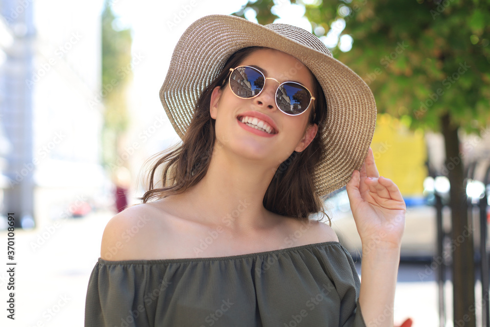 Smiling pretty girl wearing sunglasses and summer hat and looking at camera while walking in city street.