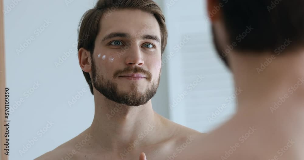 Close up mirror reflection smiling young man having fun, applying moisturizing balm on cheek after showering. Happy handsome guy using creme, enjoying morning healthcare routine alone in bathroom. Close up mirror reflection smiling young man having fun, applying moisturizing balm on cheek after showering. Happy handsome guy using creme, enjoying morning healthcare routine alone in bathroom.