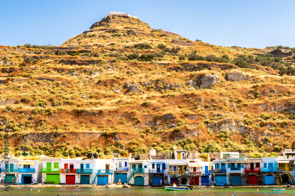 Klima fishermen village with row of traditional Greek whitewashed houses with colorful doors and windows. Grassy hill in the background, view from the sea, Milos Island, Greece.