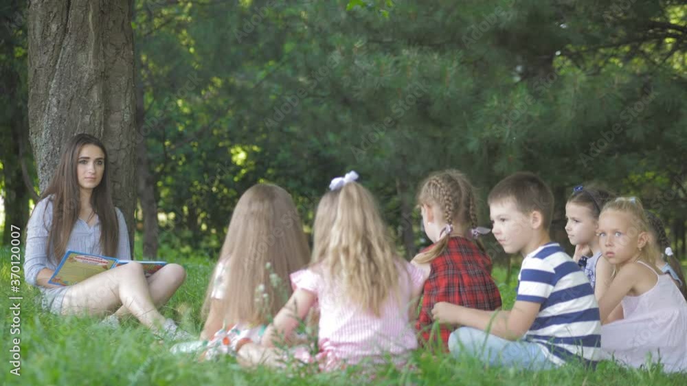 Young woman at work as educator reading book to boys and girls in park Young woman at work as educator reading book to boys and girls in park