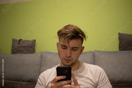 Young male with light brown hair in a white t shirt in a green room reading something off his phone AM