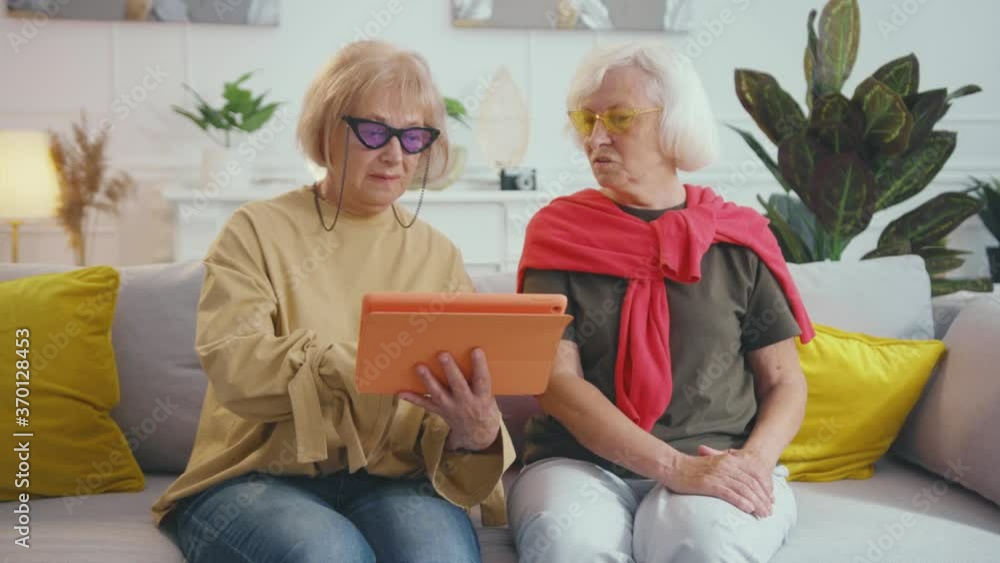 Nice beautiful and stylish old ladies using tablet computer searching moves doing e-commerce shopping from home. Concept of fashion. Generations. Elderly. Technology. Nice beautiful and stylish old ladies using tablet computer searching moves doing e-commerce shopping from home. Concept of fashion. Generations. Elderly. Technology.