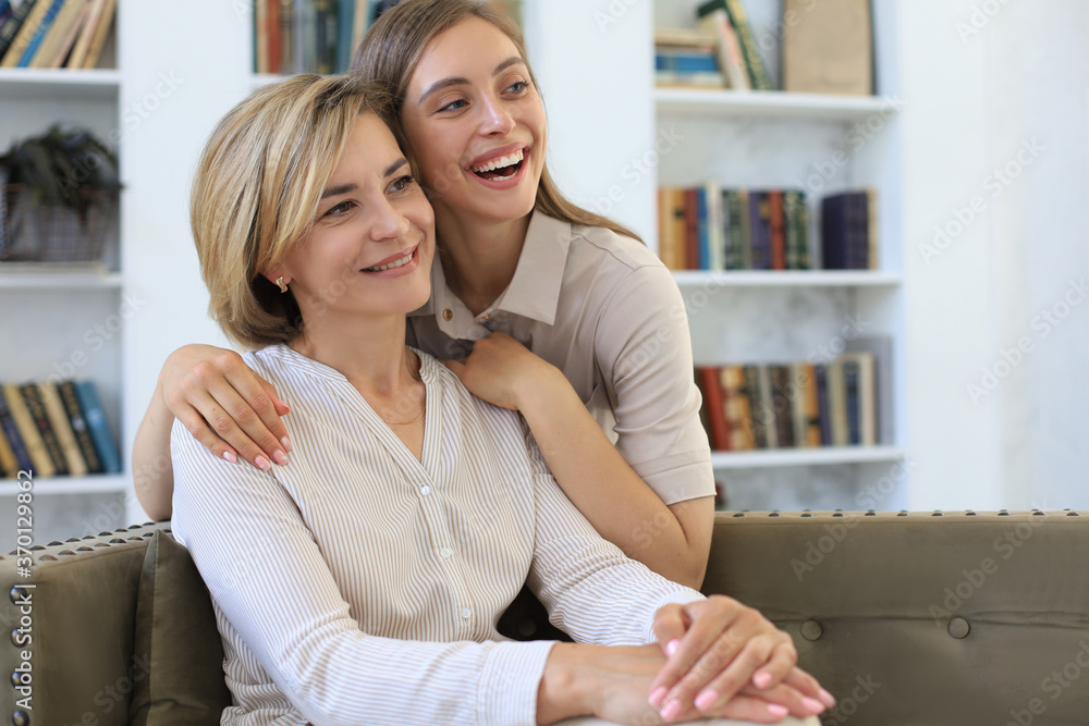 Beautiful middle aged mom and her adult daughter are hugging and smiling while sitting on couch at home. Beautiful middle aged mom and her adult daughter are hugging and smiling while sitting on couch at home.