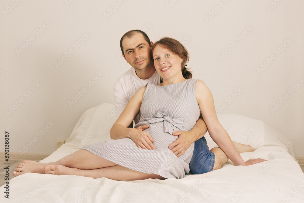 Smiling man embracing happy pregnant woman sitting on couch holding tummy with . Smiling man embracing happy pregnant woman sitting on couch holding tummy with .