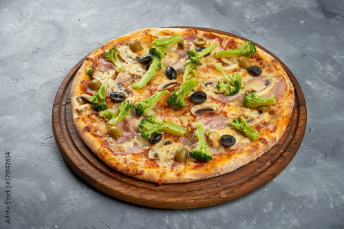 Baked italian pizza with olives, broccoli, ham and mushrooms on a wooden tray on a gray background