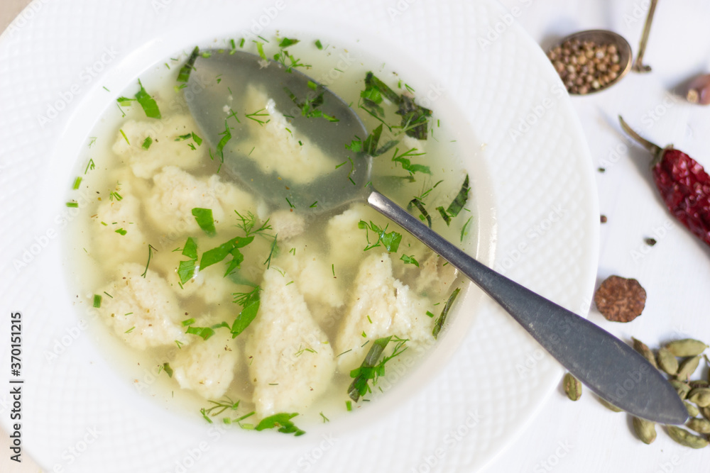 Chiken broth with dough and herbs in plate on white wooden background with spoon and spices