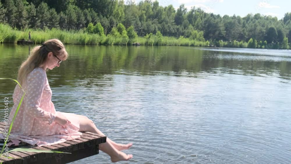 Woman on the lake. Young woman in pink dress sitting on a wooden platform on the lake and dip her feet in water, creating splashes Woman on the lake. Young woman in pink dress sitting on a wooden platform on the lake and dip her feet in water, creating splashes