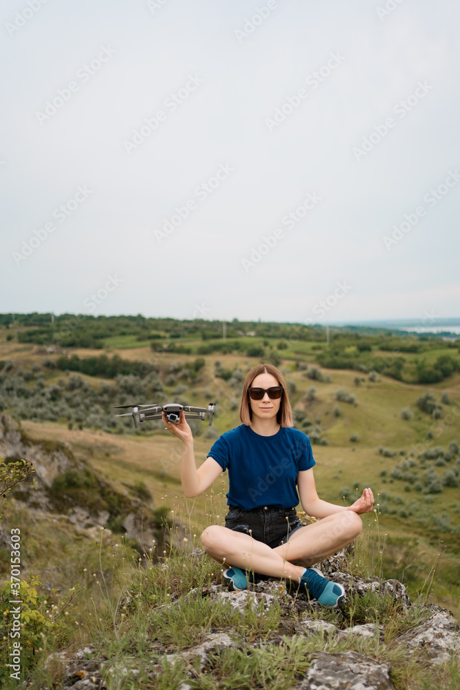 A Caucasian woman with a drone in her hand, sitting on a green rocky hill with sky in background