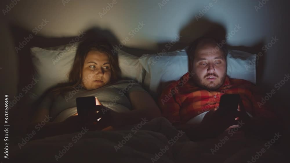 Overweight couple lying in bed and using smartphones late at night Overweight couple lying in bed and using smartphones late at night