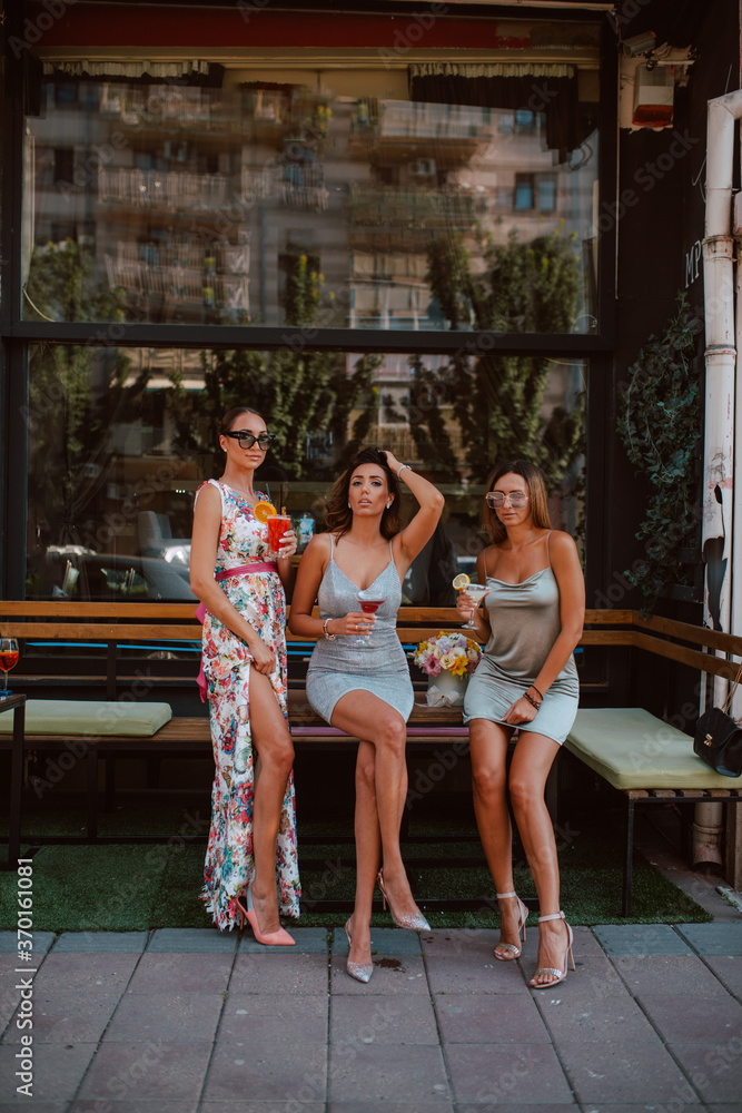 Three beautiful young caucasian women in short summer dresses are drinking cocktails in a cafe garden. Girls at party