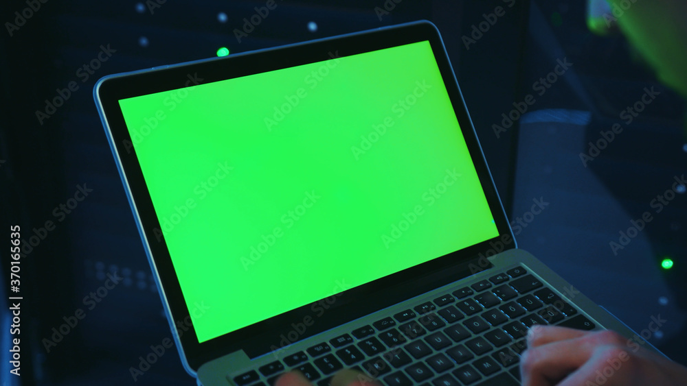 In dark data center, worker holding greenscreen mock-up laptop computer. Close-up IT technician clicking button on notebook activating modern database server cabinet. In dark data center, worker holding greenscreen mock-up laptop computer. Close-up IT technician clicking button on notebook activating modern database server cabinet.