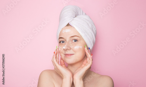 A girl with a towel on her head on a pink background and cream on her face. Hydration of the skin, skin care face, beauty.