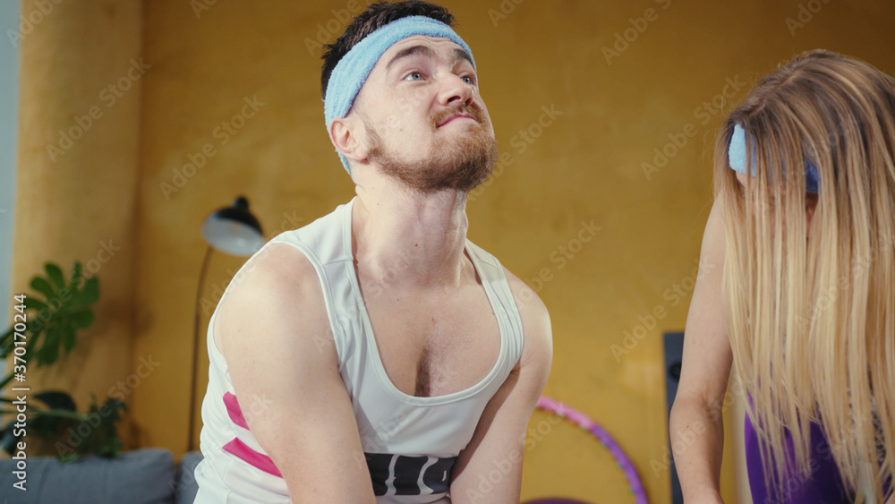 Obraz premium Weak strained bearded man lifting up heavy weight dumbbels struggling. Helpful blonde girlfriend giving support. Funny faces. Fitness parody concept.
