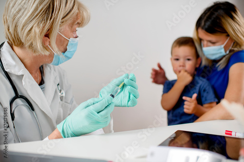 Pediatrician makes vaccination to cute Caucasian boy.Mother and doctor are wearing a protective face masks.