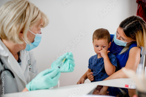 Pediatrician makes vaccination to cute Caucasian boy.Mother and doctor are wearing a protective face masks.