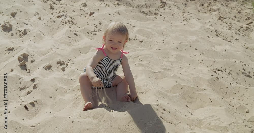Baby girl plays with sand on the beach, smiling child, hot summer time out of the city, having fun outdoors, happy childhood, cute little girl sitting on the sand