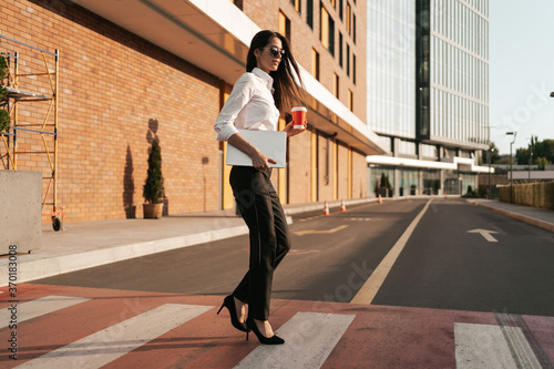 Businesswoman having a break from work and relaxing while crossing the street on zebra holding coffee to go with business center in background.