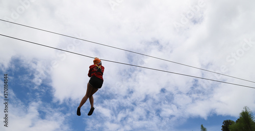 Closeup of a woman gliding on extreme trolley zip-line in adventure park with cloudy blue sky background. 