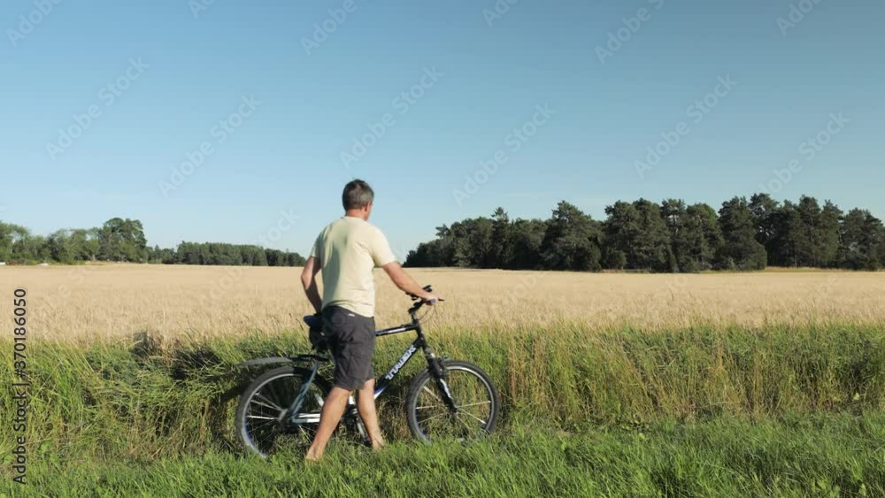 Close up view of male with bicycle going along rye field. Rye field merging with blue sky and green trees. Close up view of male with bicycle going along rye field. Rye field merging with blue sky and green trees.