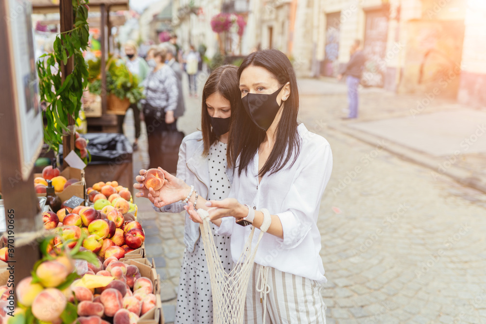 Charming woman with teenager daughter wearing a protective face mask at an outdoor fruit and veg market stall. Two females buying fresh organic peaches in farmer market, outdoors.