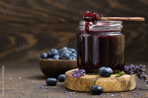 Фотография Glass jar and spoon with delicious blueberry jam with fresh berries, lavender and rosemary on brown table