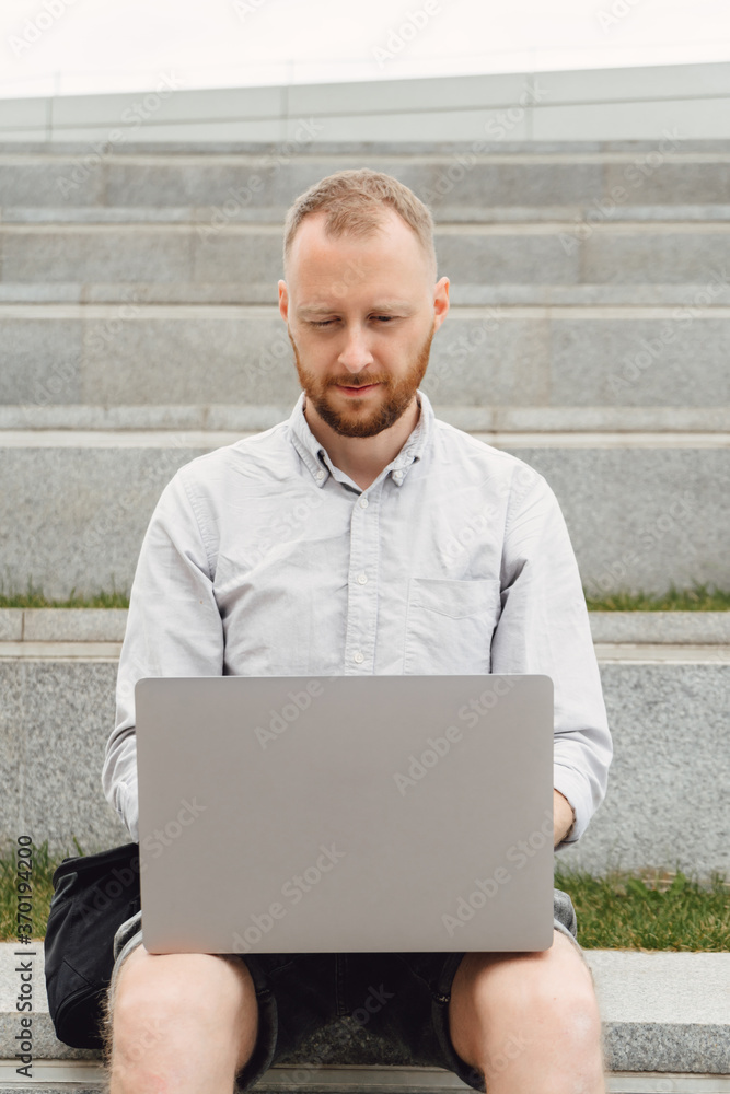 Young man works at a computer in an urban environment. Freelance programmer sits on a concrete bench in the city with a laptop. Bearded positive guy is developing new software. Young man works at a computer in an urban environment. Freelance programmer sits on a concrete bench in the city with a laptop. Bearded positive guy is developing new software.