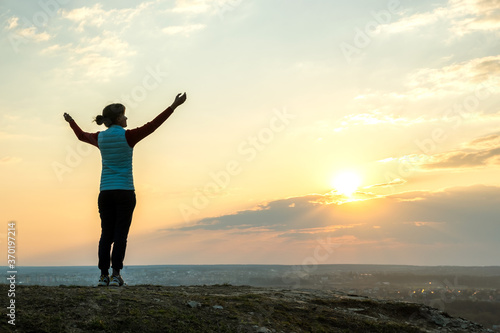 Silhouette of a woman hiker standing alone raising hands enjoying sunset outdoors. Female tourist on rural field in evening nature. Tourism, traveling and healthy lifestyle concept.