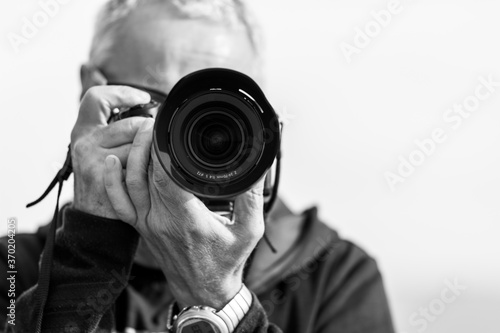 A black and white photo of a middle-aged man, in his 50th, takes a picture with a modern mirrorless camera. The camera covers his face. Blue hazy sky in the background