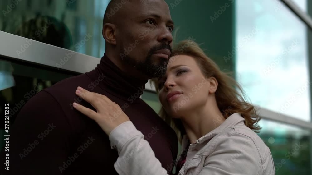 Close-up portrait of a loving couple, an African-American man and a blonde woman, they tenderly embrace against the background of the Windows of a business center on the street Close-up portrait of a loving couple, an African-American man and a blonde woman, they tenderly embrace against the background of the Windows of a business center on the street