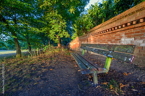 A old and broken bench in Harvington Park, Beckenham, Kent. The bench is at sunset with a red brick wall and trees behind. photo