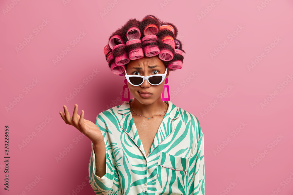 Displeased indignant woman wears curlers for getting hair styled, raises hand and puzzled by unpleasant news, wears stylish sunglasses and dressing gown, stands indoor against pink background