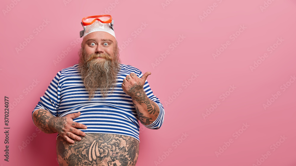 Happy bearded overweight man wears swimming goggles and cap, points finger to side, demonstrates copy space for your advertisement, keeps one hand on tattooed belly, gives instructions how to swim