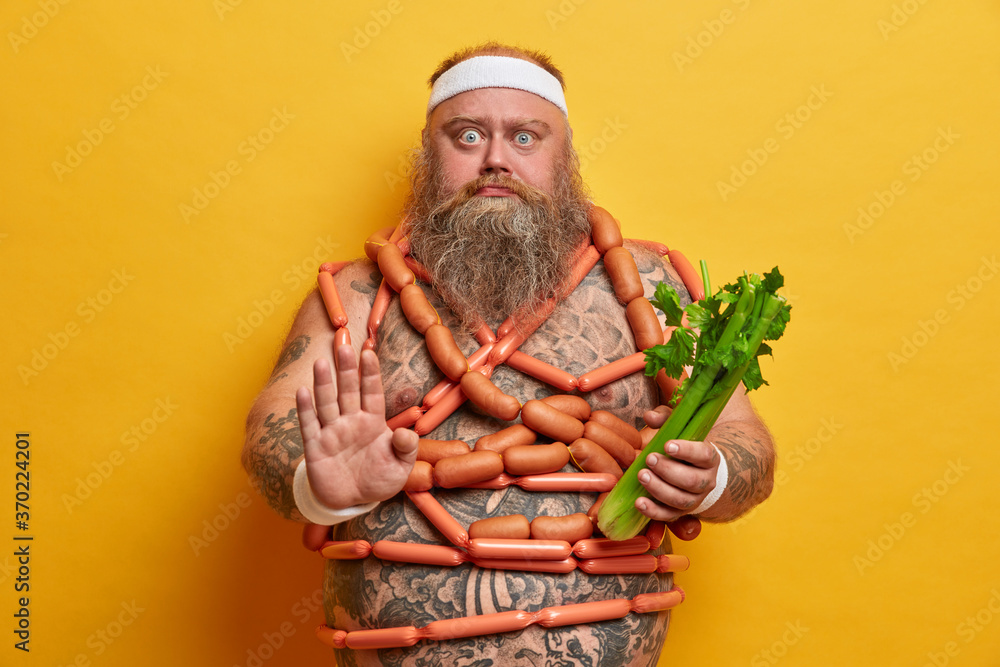 Serious motivated overweight man makes stop gesture, keeps to strict vegetarian diet and chooses celery to eat, wears sport headband, stands naked, wrapped by sausages, stands indoor. Junk food - obrazy, fototapety, plakaty 